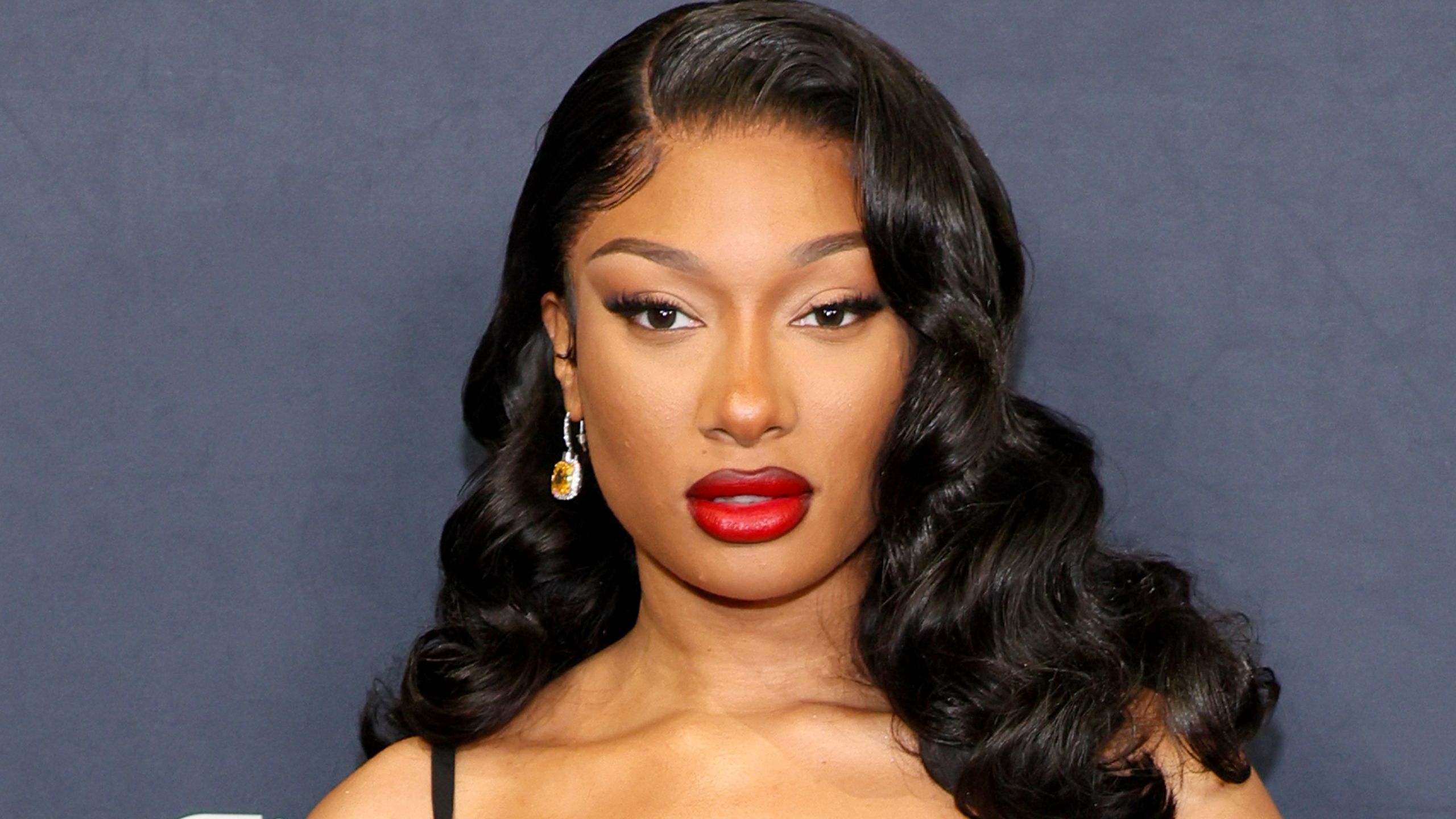 Megan Thee Stallion appears at an event in a black dress red lipstick and waves.